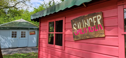 To små hytter med skilte 'SALINGER' og 'KEROUAC' ved Writers Hut på Zomerlicht, Holland, omgivet af træer.