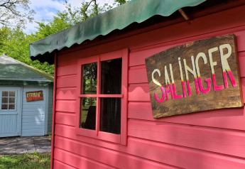 To små hytter med skilte 'SALINGER' og 'KEROUAC' ved Writers Hut på Zomerlicht, Holland, omgivet af træer.