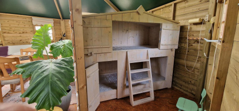 Binnenkant van een klein houten huisje met hoogslaper, eettafel, grote bladeren en kinderstoeltje.