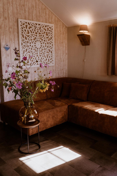 Knusse zithoek in een lodge met bruine hoekzetel, wilde bloemen in vaas en sierlijk wandpaneel.