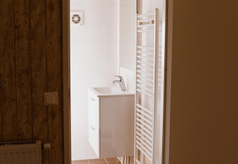 Blick in ein Badezimmer einer Lodge mit braunen Fliesen, offenem Türrahmen, Waschbecken und Handtuchheizkörper.