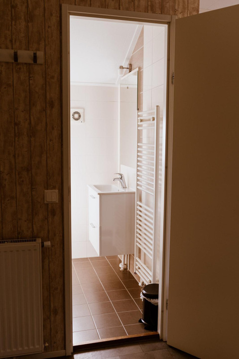 Blik in een lodgebadkamer met open deur, bruine tegels, witte wastafel en handdoekradiator.