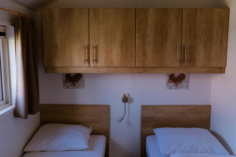 Deux lits simples sous des placards en bois dans une chambre de lodge, près d'une fenêtre à rideaux.