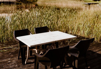 Terrasse extérieure avec chaises noires en plastique et table blanche au bord d’un lac, entourée de lodges.