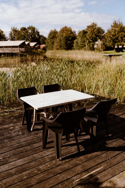 Terrasse extérieure avec chaises noires en plastique et table blanche au bord d’un lac, entourée de lodges.