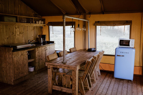 Cucina glamping rustica con tavolo in legno, sedie, lampade sospese, frigorifero blu e microonde in tenda.