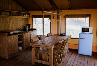 Cocina glamping rústica con mesa de madera, sillas, luces colgantes, nevera azul y microondas en tienda.