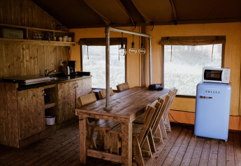 Cucina glamping rustica con tavolo in legno, sedie, lampade sospese, frigorifero blu e microonde in tenda.