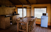 Cucina glamping rustica con tavolo in legno, sedie, lampade sospese, frigorifero blu e microonde in tenda.
