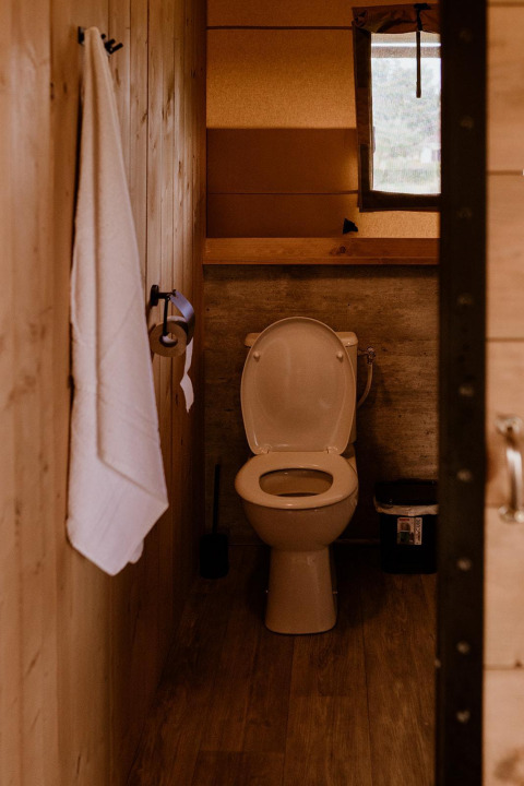 Baño de tienda safari con paredes de madera, inodoro, toalla blanca, ventana y papel higiénico.