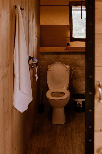Safari tent bathroom with wooden walls, toilet, tissue roll, white towel, window and trash can.