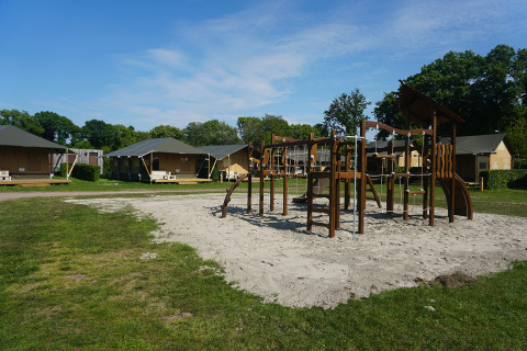 Playground with climbing equipment in front of several safari tents at a campsite under a blue sky.
