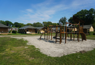 Parque infantil con estructura para trepar frente a varias tiendas safari en un camping bajo cielo azul.
