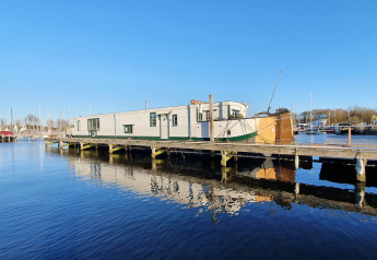 Houseboat De Zwerver docked at Marina Parcs Naarden in the Netherlands, reflected in calm blue water.