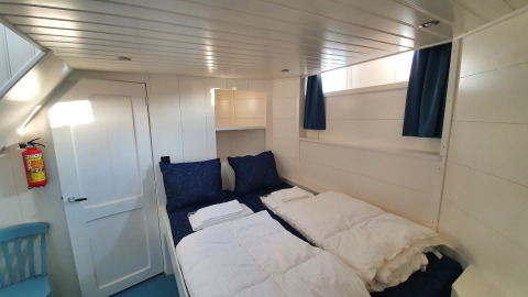 Lichte slaapkamer met tweepersoonsbed, blauw beddengoed en ramen op woonboot De Zwerver in Nederland.