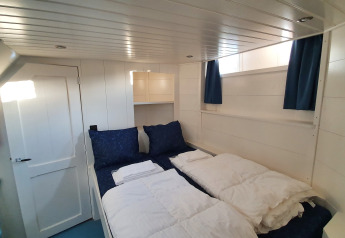 Lichte slaapkamer met dubbel bed, blauw beddengoed en ramen op woonboot De Zwerver in Nederland.