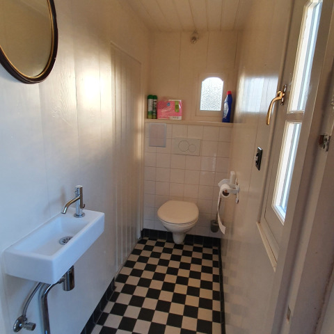 Smal toilet op een woonboot met zwart-wit geblokte vloer, zwevend toilet, wastafel en raam.
