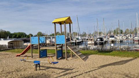 Parque infantil con tobogán y balancín sobre arena junto a un puerto deportivo y glamping en verano.