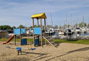 Playground with slide and seesaw on sand, next to a marina and tents at a glamping accommodation.