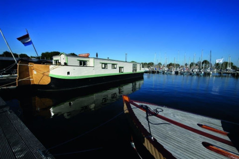 Woonboot De Zwerver aangemeerd in Marina Parcs Naarden, Nederland, onder een stralende blauwe lucht.