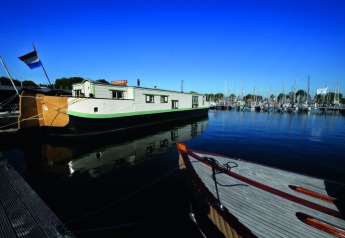 Casa flotante De Zwerver atracada en Marina Parcs Naarden en los Países Bajos bajo un cielo azul brillante.