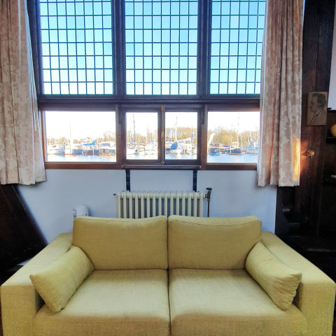 Sala de una casa flotante con sofá amarillo, grandes ventanas y vista al puerto deportivo con barcos.