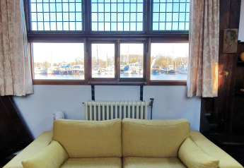 Woonkamer op een woonboot met gele bank, grote ramen en uitzicht op een jachthaven met boten.