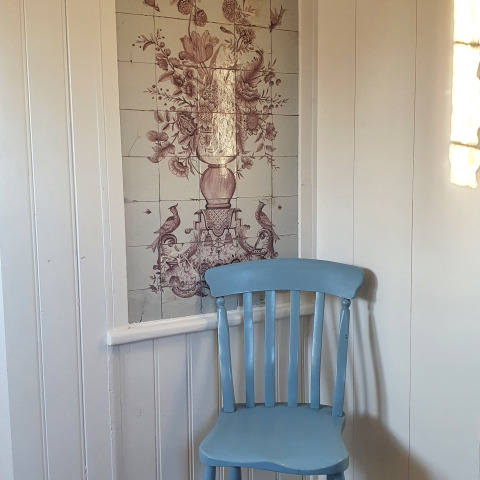 Una foto de una silla azul claro frente a paneles blancos y azulejos decorativos de flores y pájaros en Holanda.