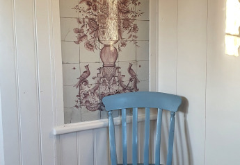 Et billede af en lyseblå stol foran vægpaneler og vægfliser med blomster- og fugledesign i Holland.