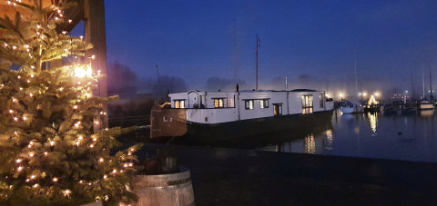 Aftenbillede af Houseboat De Zwerver i Marina Parcs Naarden, oplyst juletræ og både i baggrunden.
