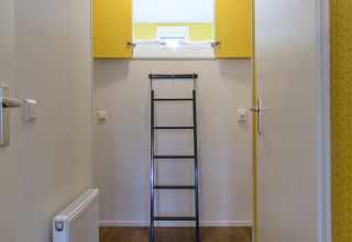 Pasillo en una cabaña moderna con una escalera negra hacia una cama elevada, puertas amarillas y suelo de madera.