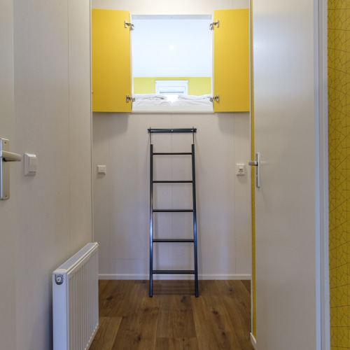 Pasillo en una cabaña moderna con una escalera negra hacia una cama elevada, puertas amarillas y suelo de madera.