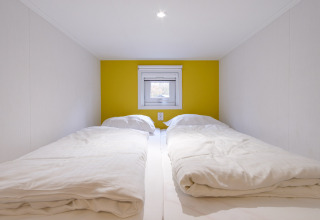 Dos camas individuales con edredones blancos en una cabaña compacta con pared amarilla y ventana pequeña.