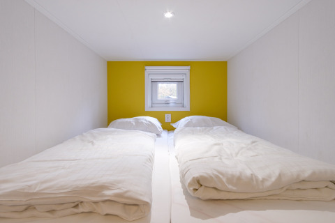 Dos camas individuales con edredones blancos en una cabaña compacta con pared amarilla y ventana pequeña.