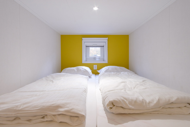 Twee eenpersoonsbedden met witte dekbedden in een compacte lodge met gele muur en klein raam.