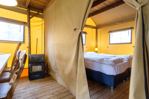 Vista interior de una tienda safari Verandalodge con cama, sillas de madera y estufa en Kampeerdorp de Zandstuve.