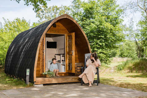 Eine Frau und ein Kind entspannen vor einer Holzhütte im Pod-Stil bei Kampeerdorp de Zandstuve, Niederlande.