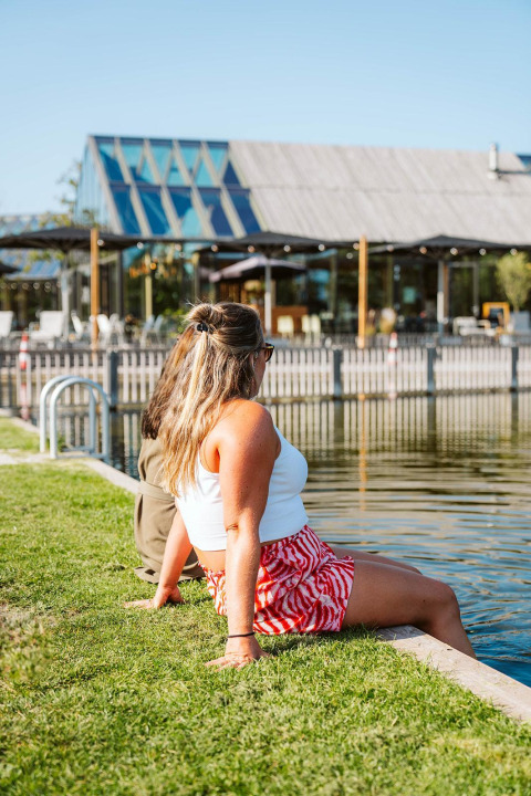 Due persone si rilassano vicino all'acqua davanti a una moderna struttura glamping con facciata in vetro in una giornata di sole.
