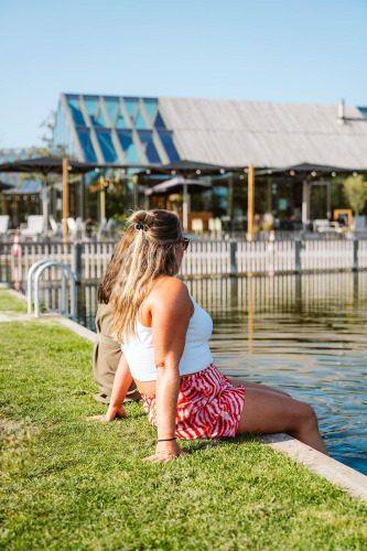 Twee mensen ontspannen aan het water voor een moderne glamping-accommodatie met glazen gevel op een zonnige dag.