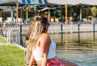 Dos personas se relajan junto al agua frente a un alojamiento glamping moderno con fachada de cristal en día soleado.