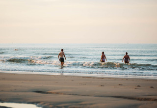 Mensen lopen het water in op een rustige strand bij een glamping-accommodatie in de avondzon.