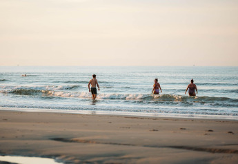 Des personnes marchent dans les vagues sur une plage paisible près d’un site de glamping en soirée.