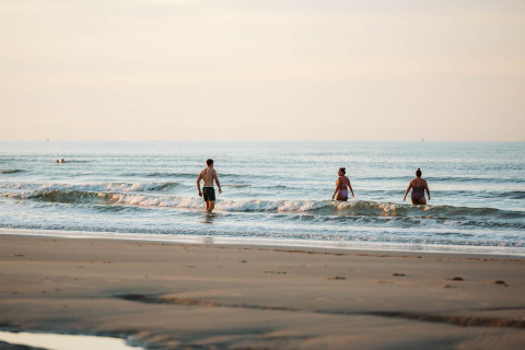 Des personnes marchent dans les vagues sur une plage paisible près d’un site de glamping en soirée.