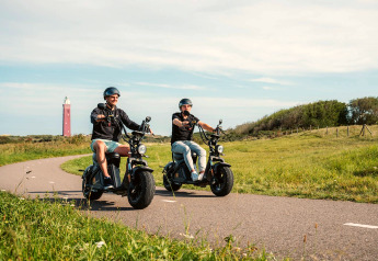 Two people riding electric scooters on a winding path by a glamping site, with a lighthouse in the distance.