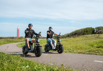 Twee mensen rijden op elektrische scooters over een pad bij een glamping, met een vuurtoren in de verte.