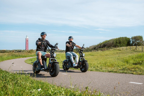 Twee mensen rijden op elektrische scooters over een pad bij een glamping, met een vuurtoren in de verte.