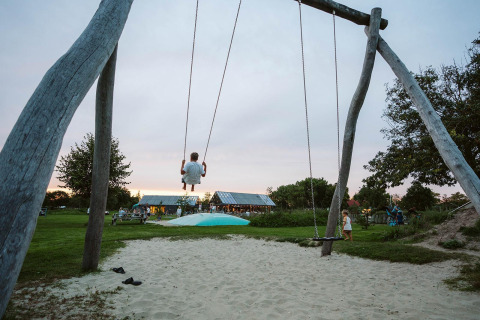 Een kind schommelt hoog op een grote houten schommel bij een glampingaccommodatie met zand en gras.