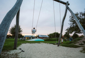 Een kind schommelt hoog op een grote houten schommel bij een glampingaccommodatie met zand en gras.