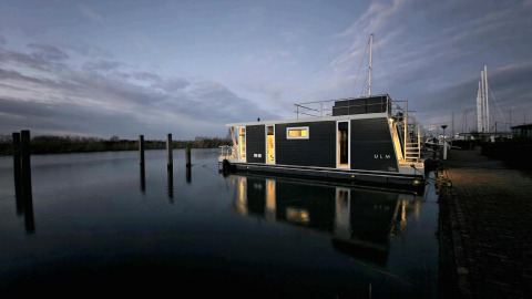 Casa flotante Havenlodge Lelystad en Marina Parcs Lelystad, Países Bajos, iluminada al anochecer sobre agua tranquila.