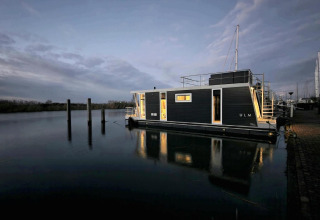 Casa flotante Havenlodge Lelystad en Marina Parcs Lelystad, Países Bajos, iluminada al anochecer sobre agua tranquila.